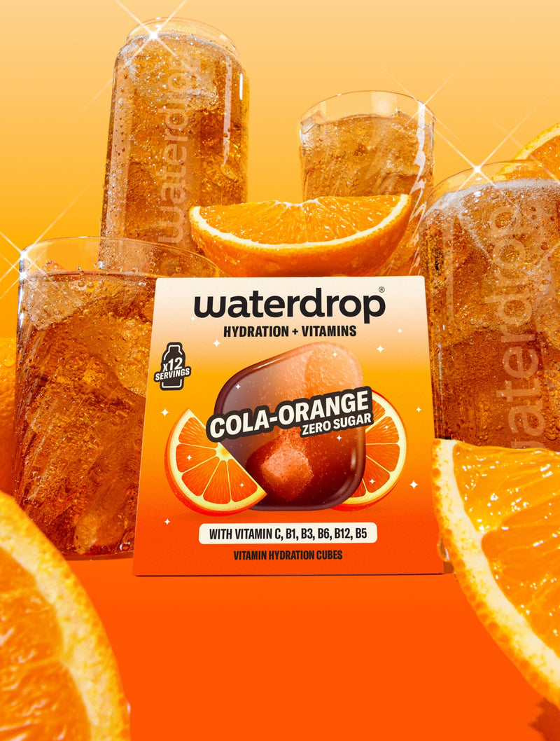 Krabička příchuti Cola Orange s 12 ks umístěná uprostřed, v pozadí jsou skleničky waterdrop® naplněné nápojem a obrázek doplňují plátky pomeranče.
