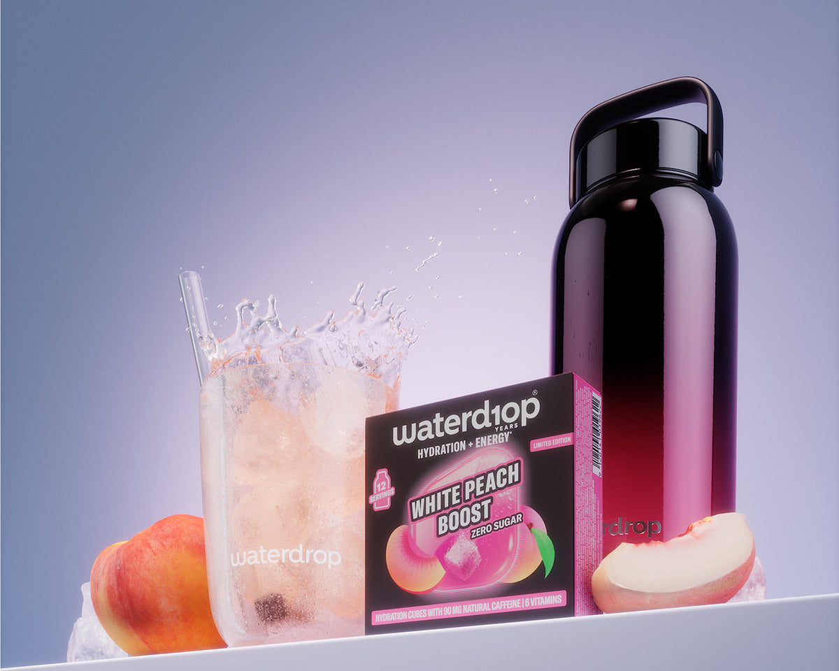 Krabice produktu White Peach Boost vedle sklenice s ledem a černé opakovatelné láhvi na bílem regálu.