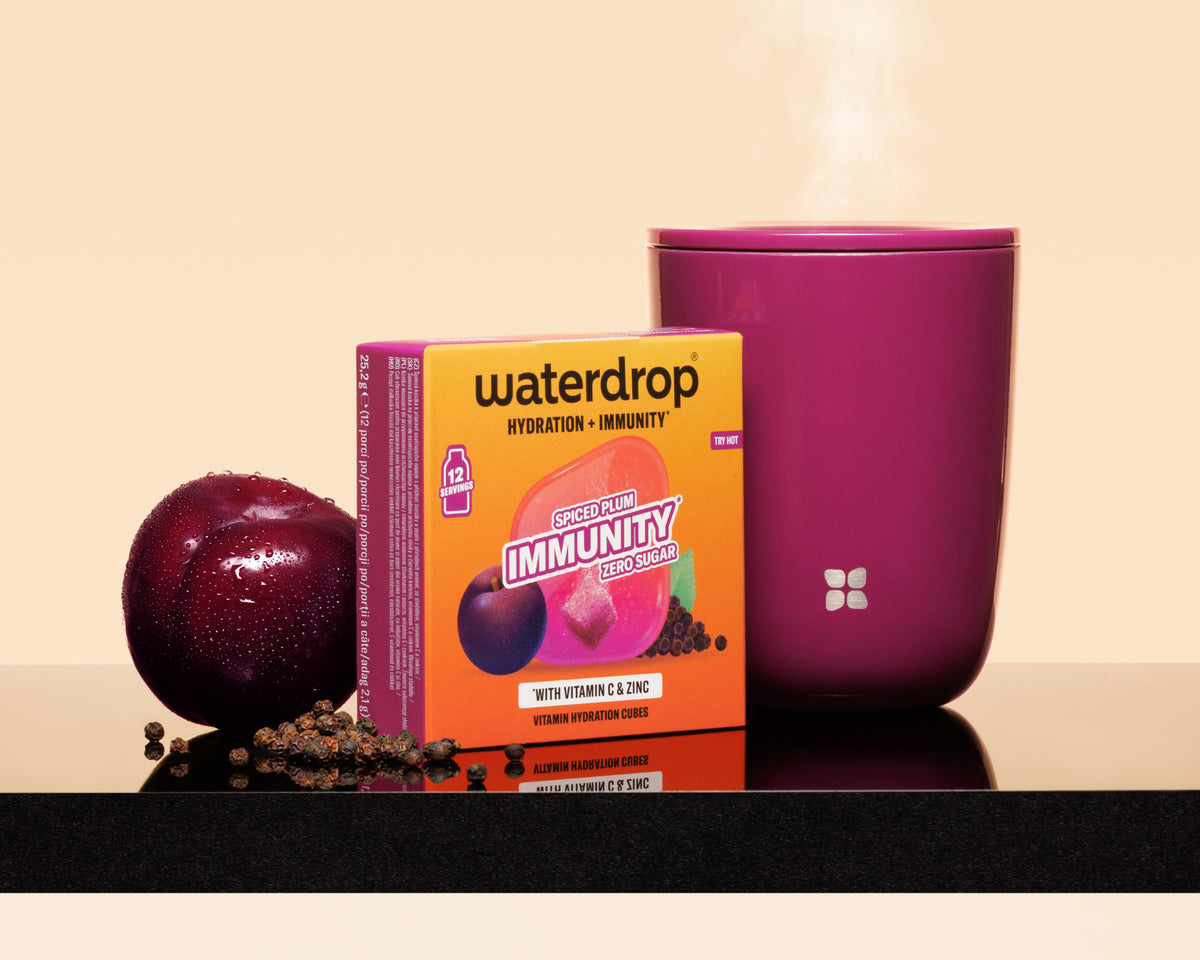 Fialová šálek s parou vedle oranžové krabice Waterdrop, švestka a posypané semena na lesklé ploše.